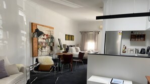 A modern hotel room with a dining area, a sofa, a chair, a small table, and a kitchen counter with amenities.