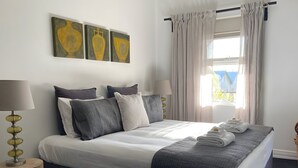 A neatly made bed with a grey and white duvet, a bedside table with a lamp, and a window with curtains.