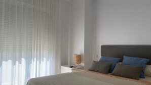 Een slaapkamer met een bed, nachtkastje, lamp en dunne gordijnen.