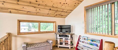 Una habitació acollidora amb sostre de fusta, una cadira amb coixí, un moble televisió ple de llibres i una finestra amb persianes.