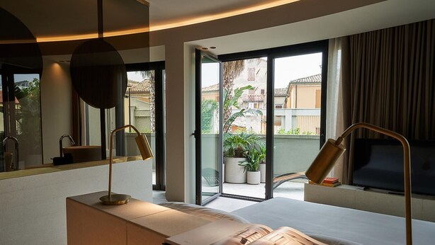 Una habitación de hotel moderna con una cama grande, una mesita de noche con una revista, un escritorio con una lámpara, un ventanal con vistas a edificios y plantas, y una puerta corrediza al balcón.