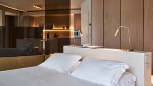 Una habitación de hotel moderna con una cama grande, una mesita de noche con lámpara, y vistas al baño y a la cocina a través de un separador de vidrio.