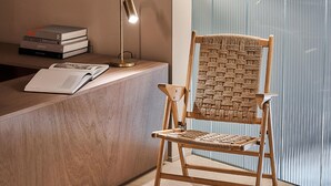 Ein Holzstuhl mit gewebter Rückenlehne und Sitzfläche, vor einem Empfangstresen mit Lampe und Büchern platziert.