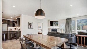Een moderne keuken met donkere kastjes, een houten eettafel en barkrukken. Er is een open haard met een televisie erboven, en een groot raam met uitzicht op een bos.