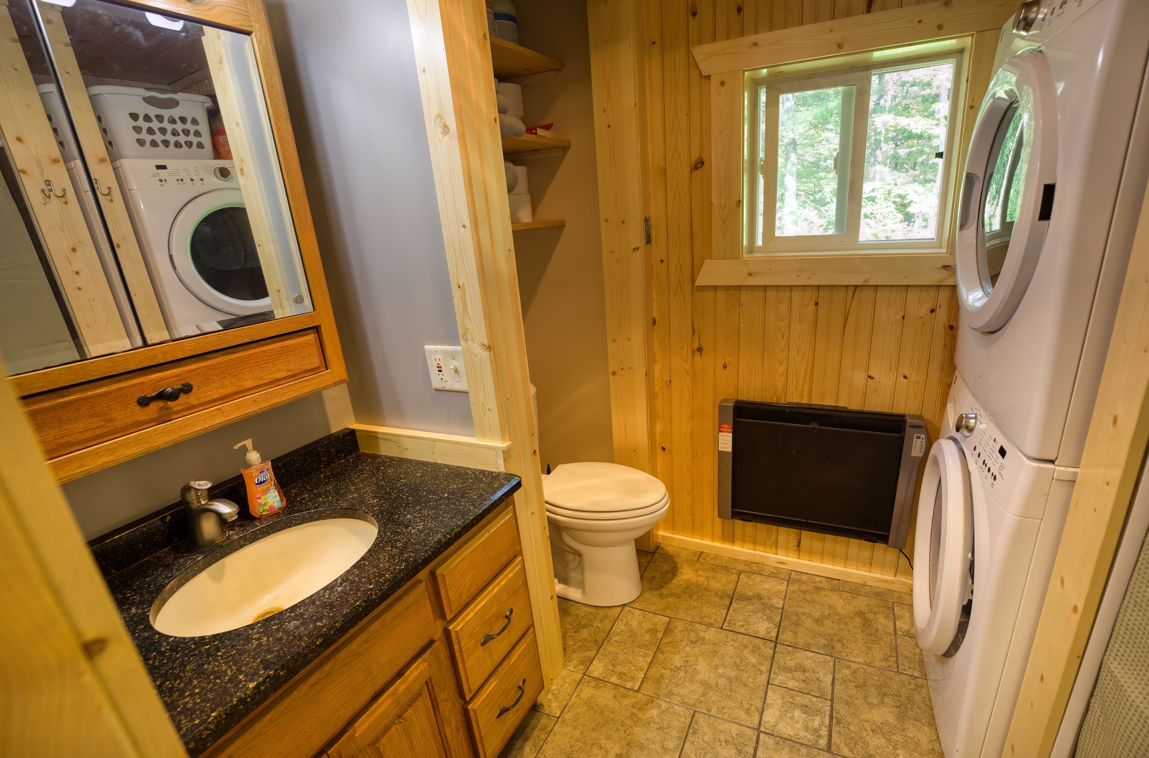 A compact bathroom with a sink, toilet, and washing machine, wooden walls, and a window.