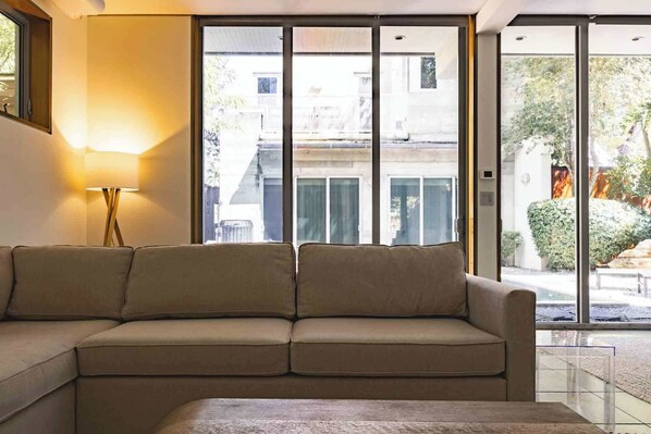 Ein Wohnzimmer mit einem beigen Sofa, einer Stehlampe und großen Fenstern mit Blick ins Freie.