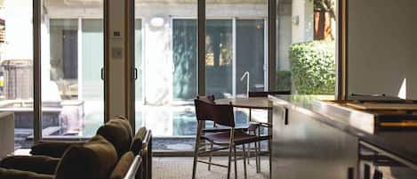 A room with a sofa, a table, and chairs near large windows with a view of a building and greenery outside.