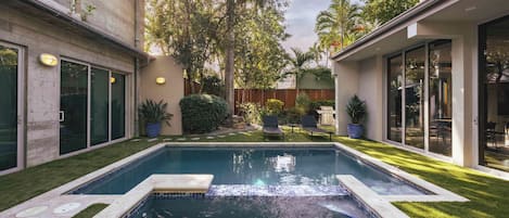 Una piscina con borde de piedra, rodeada por un césped cuidado y plantas en macetas.