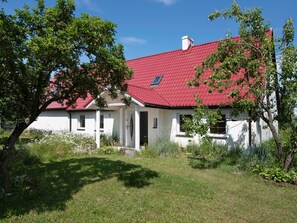 Una casa blanca amb teulada vermella, un petit porxo i un jardí amb diverses plantes i un arbre.