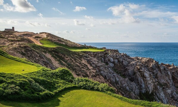 A coastal golf course with a rocky cliff, a bunker, and a flag on the green.