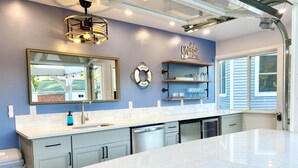 A modern kitchen with a large island, a built-in dishwasher, and a wall-mounted sink. There are shelves above the sink holding glassware and a 'Welcome' sign. - Beautiful cottage with lake access and bonus entertaining space! (Lakeview)