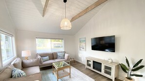 A spacious living room with a high ceiling, a large window, a sofa set, a coffee table, a TV, and a plant. - Beautiful cottage with lake access and bonus entertaining space! (Lakeview)