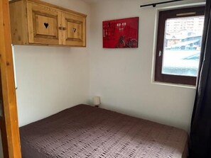 A bedroom with a wooden wardrobe, a red poster, a window with a view of buildings, and a bed with a textured cover.