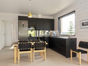 A modern kitchen with black cabinets, a stainless steel refrigerator, a dining area with wooden chairs, and a large window providing a view of the outdoors. - Villa horihane by Interhome (Inari)