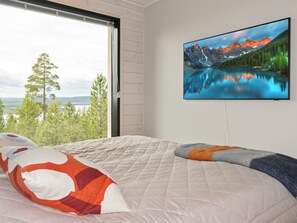 Makuuhuoneessa on suuri ikkuna, sängyssä on peitto ja seinällä on televisio, josta näkyy vuoristomaisema.