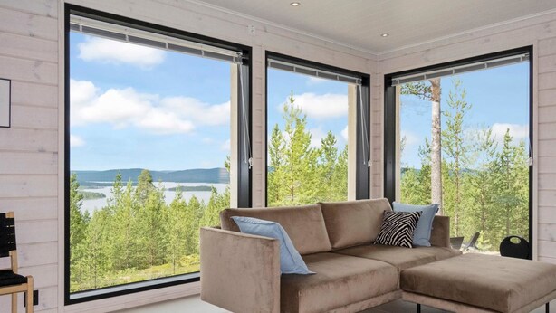 Un salon moderne avec un canapé d’angle beige, de grandes fenêtres donnant sur un lac et une forêt, et un design minimaliste.