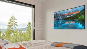 Une chambre avec une grande fenêtre, un lit avec une couette, un oreiller et un téléviseur fixé au mur qui diffuse un paysage de montagne.