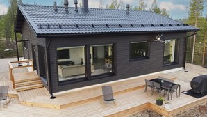 Une cabane moderne aux couleurs sombres, dotée d’un toit en pente, de grandes fenêtres, d’une terrasse en bois et de mobilier d’extérieur.