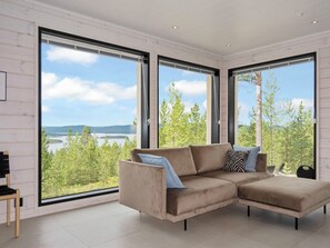 A modern living room with a beige sectional sofa, large windows, and a view of a lake and forest. - Villa horihane by Interhome (Inari)