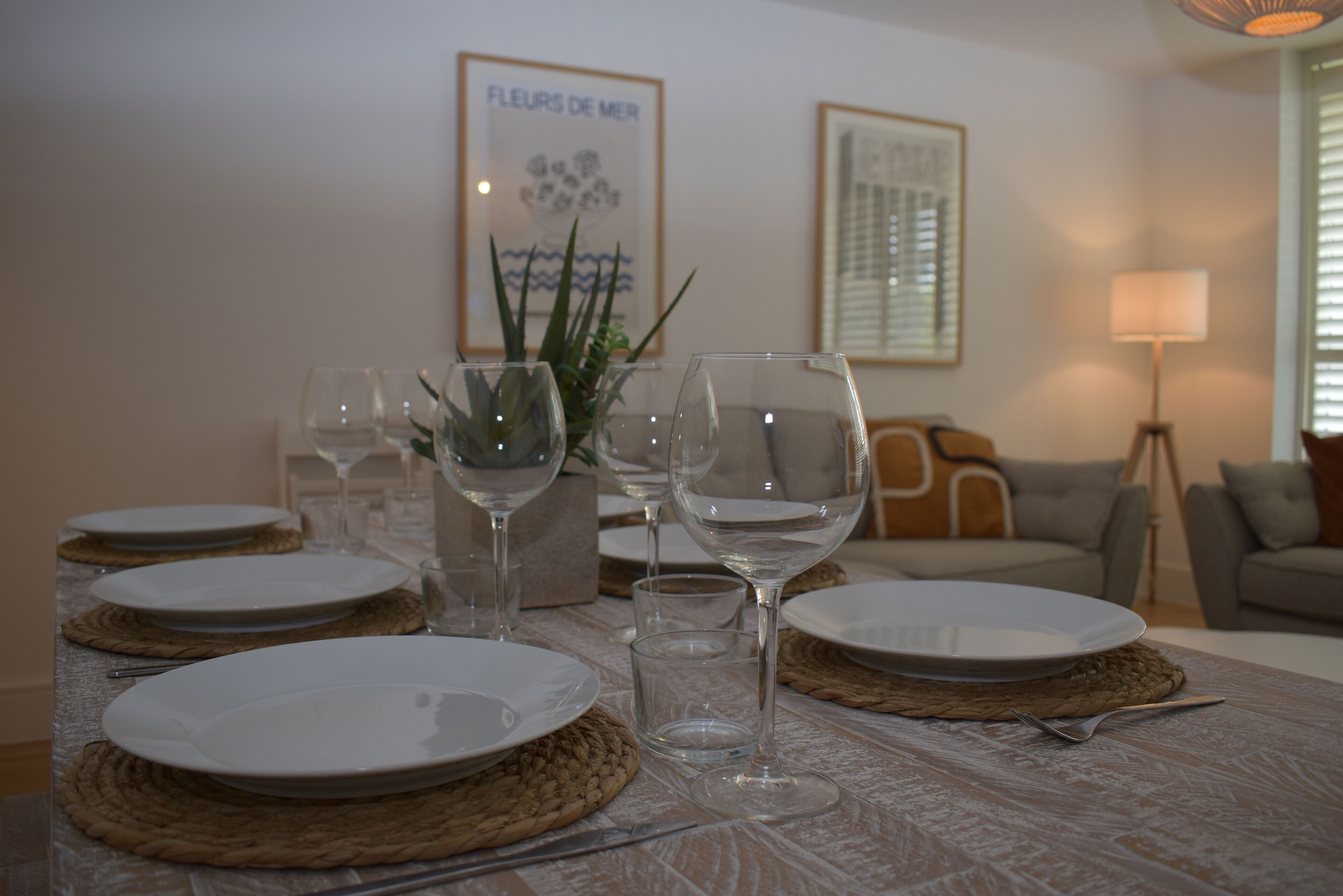 A dining table set with plates, glasses, and cutlery, with a plant and framed artwork in the background.