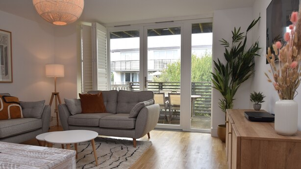 A modern living room with a grey sofa, a white coffee table, and a wooden cabinet. There is a large window with a view of a balcony and a building. - Les Dunes (Rye)