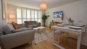 A modern living room with a grey sofa, a dining table set for four, a large window with blinds, a hanging light fixture, and a potted plant. - Les Dunes (Rye)