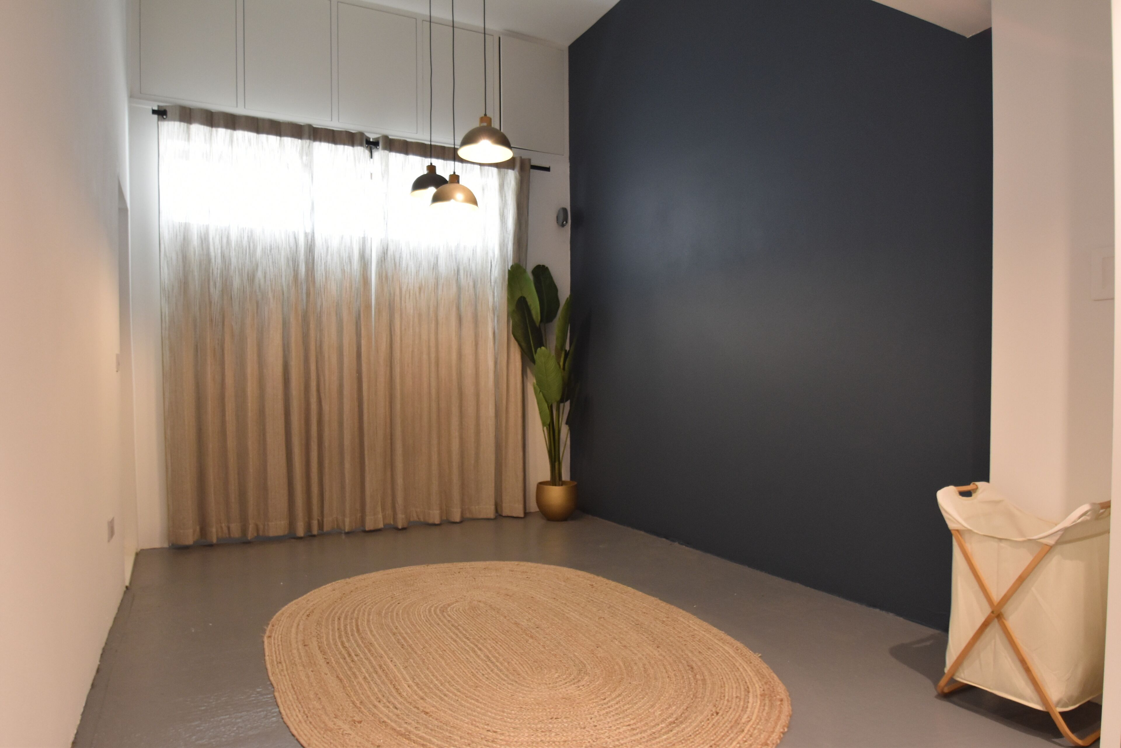 A room with a dark wall, a large window with sheer curtains, a woven rug, a potted plant, and a white chair.