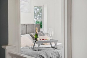 Une chambre à coucher avec un lit, une table de chevet sur laquelle se trouvent une bouteille et des verres, et une fenêtre donnant sur un paysage verdoyant.