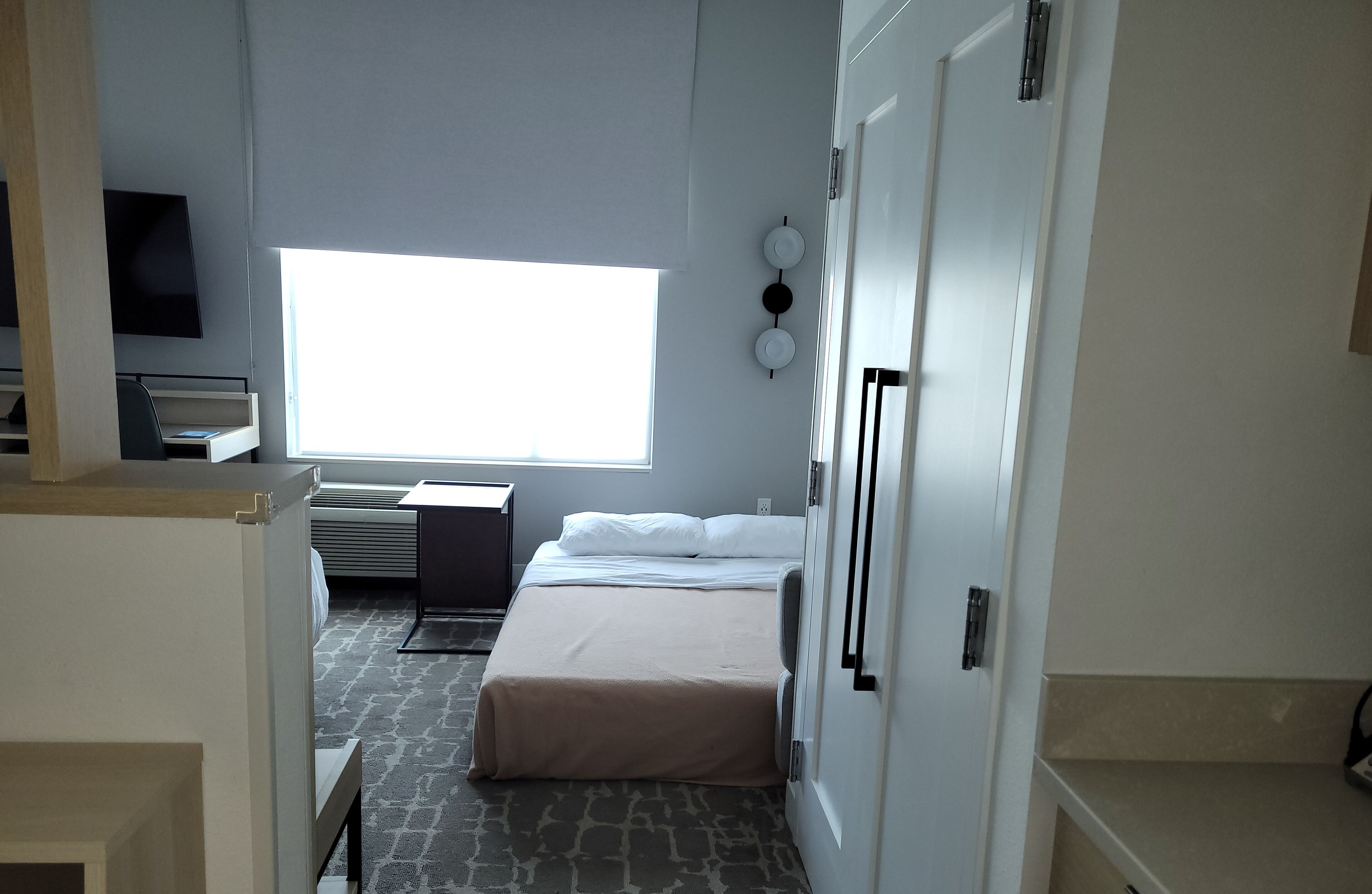 A hotel room with a bed, a desk, a chair, a heater, and a window.