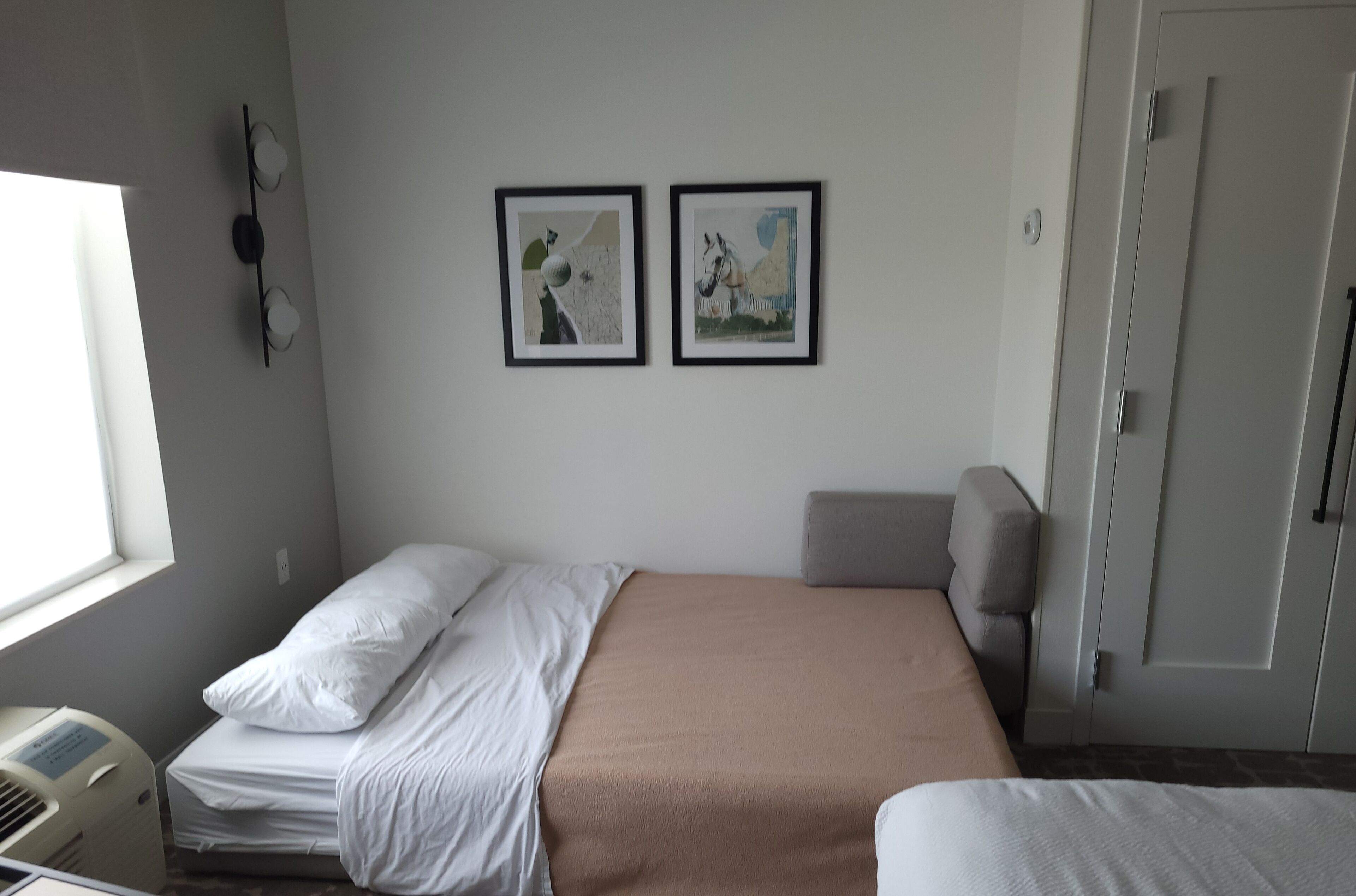 A bedroom with a bed, a bedside table, a heater, a window, and two framed pictures on the wall.