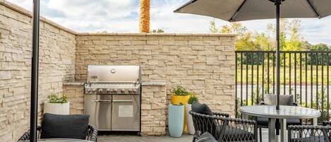 An outdoor patio with a stone wall, a grill, wicker chairs, and a round table.
