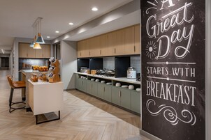 A modern kitchen with wooden cabinets, a central island, and a chalkboard with 'A Great Day Starts with Breakfast'.