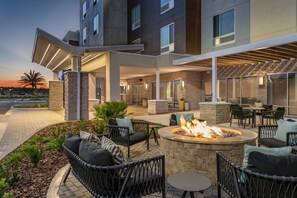 Un patio exterior moderno con mobiliario de mimbre, una fogata central y una chimenea de piedra.