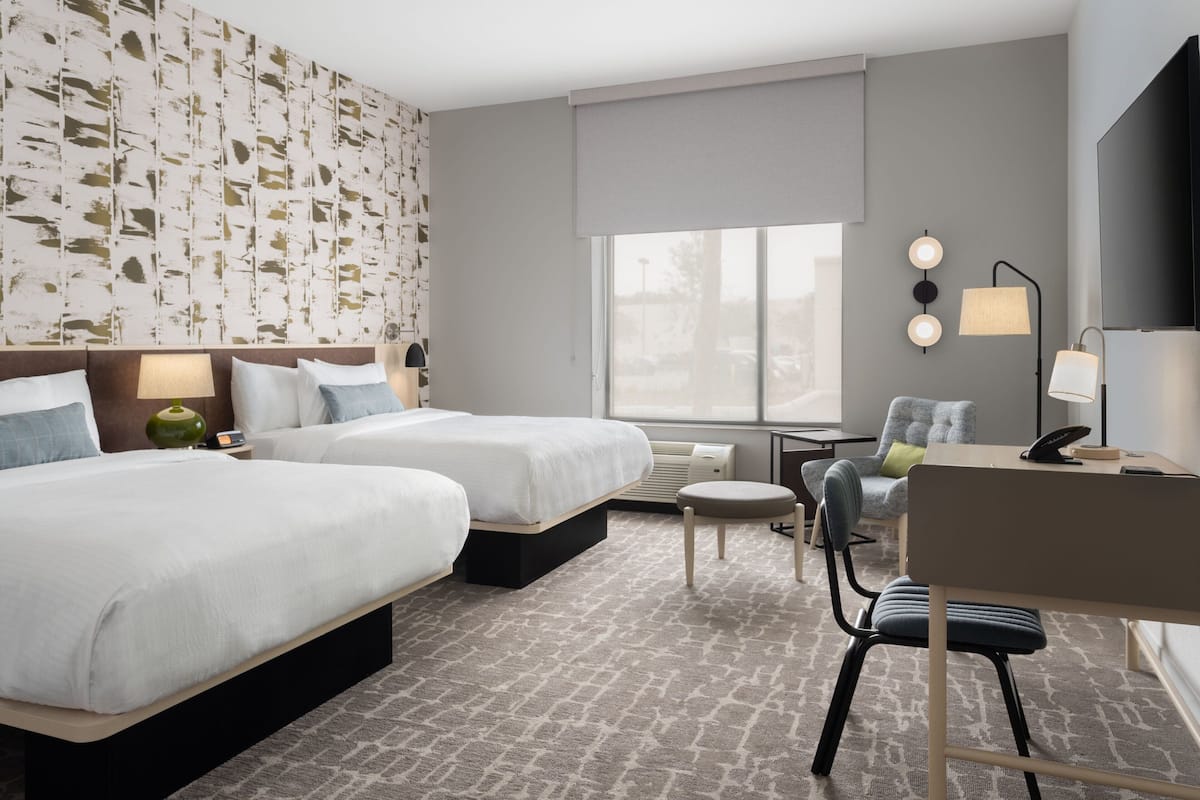 A hotel room with two beds, a desk, a chair, a small table, and a large window.