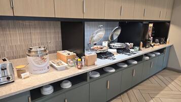 En buffet med forskellige madretter, herunder en stor skål havregryn, en kaffemaskine og flere serveringsfade.