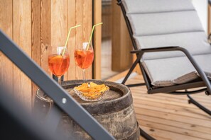 Deux verres d’une boisson rouge, chacun muni d’une paille, posés sur un tonneau en bois, un bol de chips et un fauteuil de relaxation.