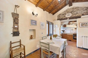 A rustic dining area with a wooden table set for two, a stone wall, and a kitchen in the background. - Vacation Home 'Casa Vittorina' with Garden, Terrace & Wi-Fi (Cesio)