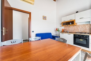 Un comedor con mesa y sillas de madera, sofá azul, armarios blancos, salpicadero de azulejos y horno empotrado.