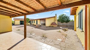 Un'area patio coperta con pavimento piastrellato, una pergola in legno e un edificio giallo con un camino in mattoni.