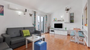 Una sala moderna con un sofá gris, una mesa de centro blanca, un televisor de pantalla plana, una pequeña zona de cocina con encimera blanca y un ventanal corredero que da a un espacio exterior.