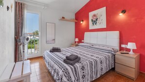 Un dormitorio con cama, mesitas de noche, ventilador, ventana con cortinas, pared roja y vistas al balcón.