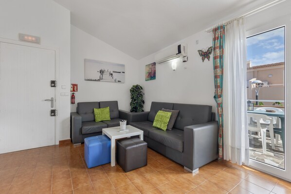Un salon moderne avec un ensemble de canapés gris, une table basse blanche et un pouf bleu. Une porte-fenêtre coulissante donne accès à un espace extérieur.