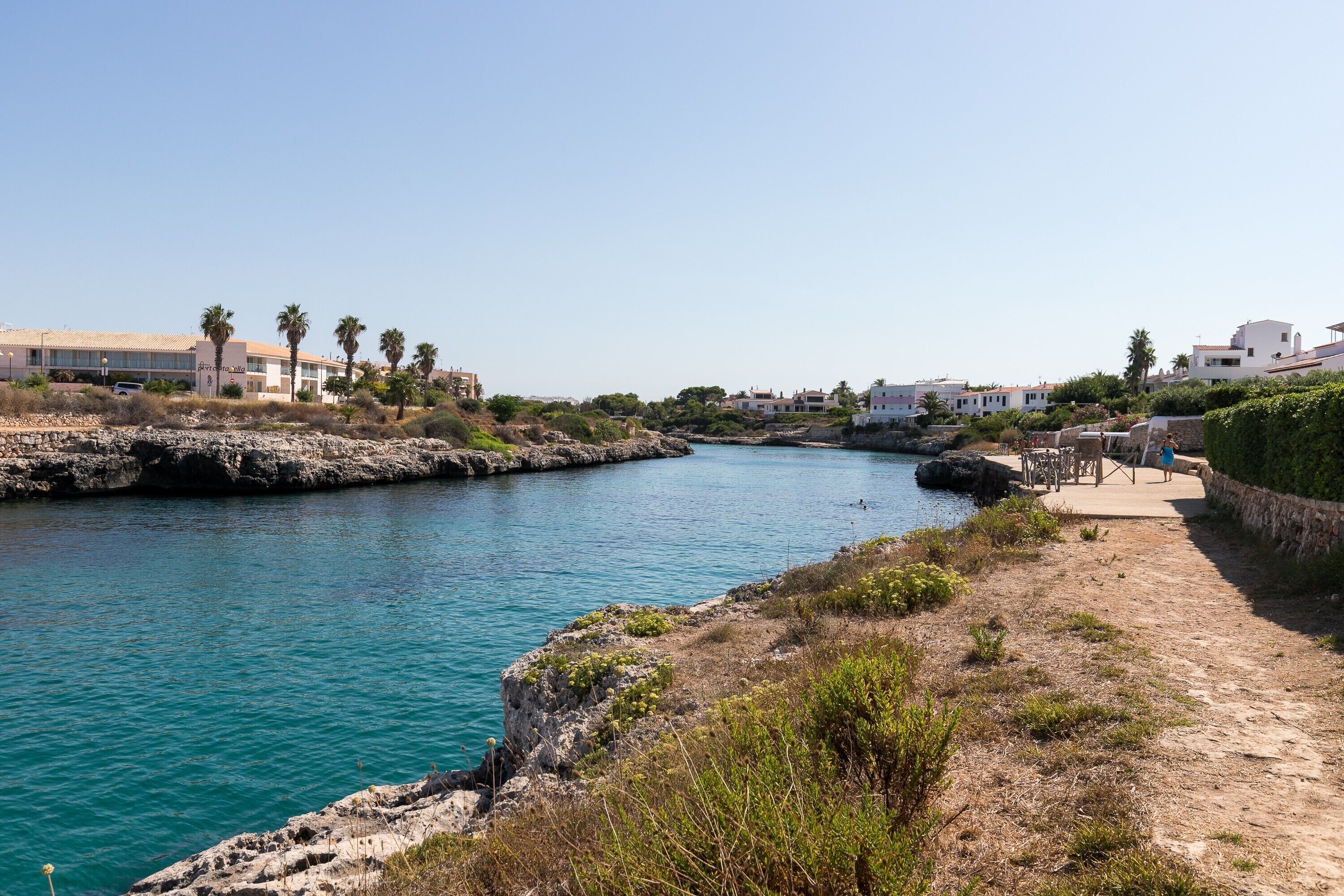 Una zona costera con mar azul cristalino, un litoral rocoso y edificios al fondo.