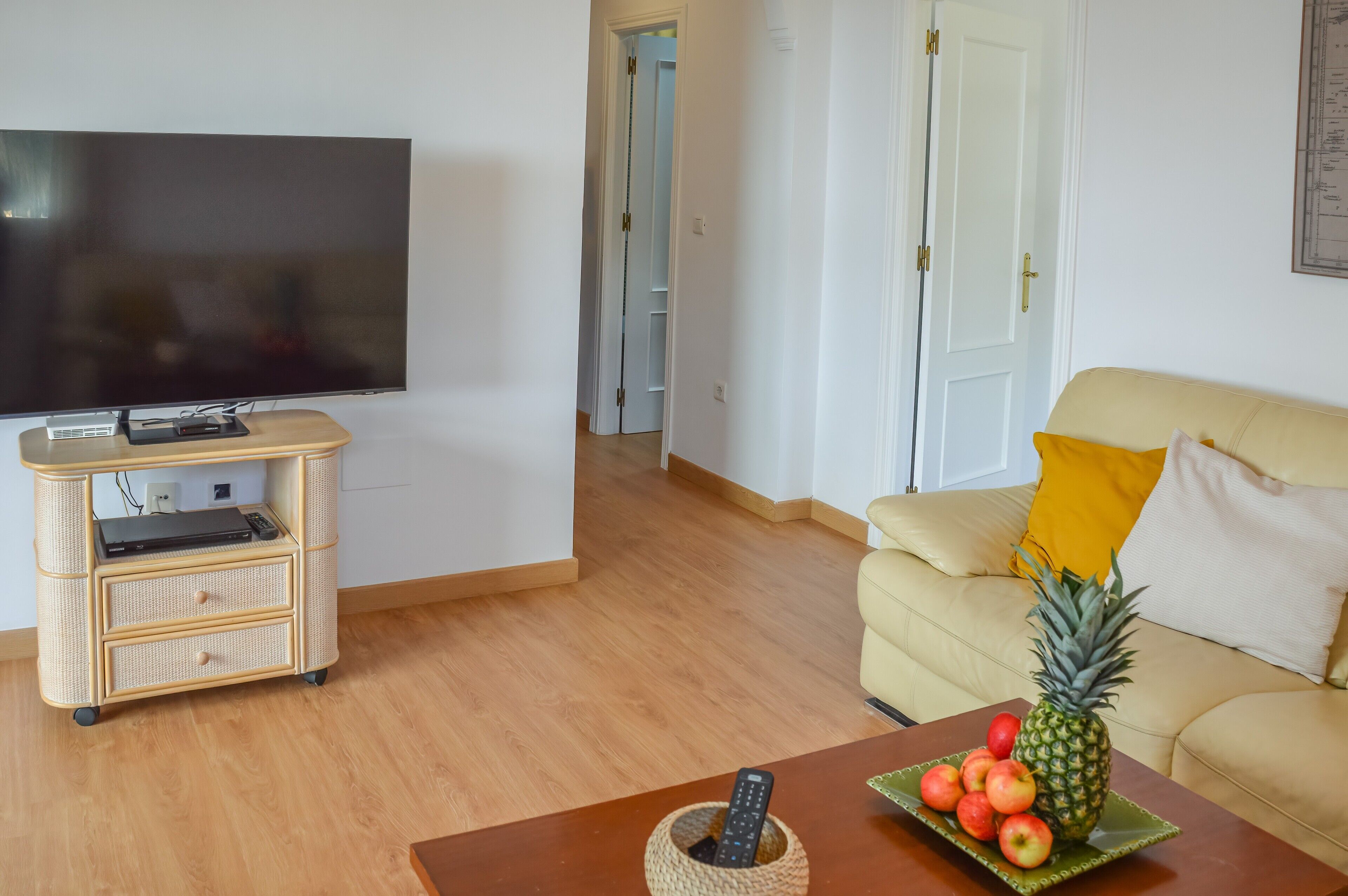 Una sala de estar con televisor, un sofá, una mesa de centro con fruta y un mueble de almacenamiento de mimbre.