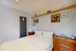 A bedroom with a bed, a wooden dresser, a nightstand, and a shelf with books and pictures.