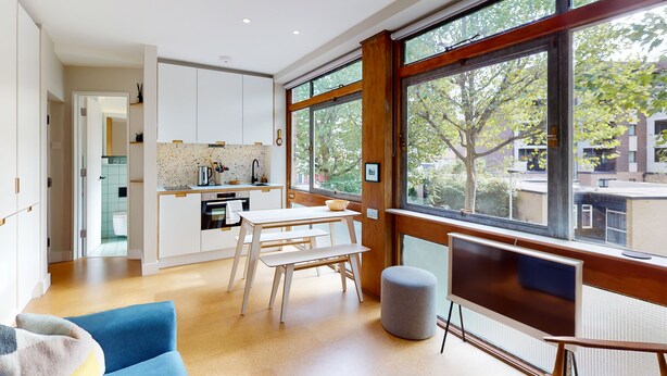 A modern kitchen with white cabinets, a dining area with a blue sofa, and large windows offering a view of trees and buildings. - UK.LO.DCR - Spacious 1 bedroom apartment in Kennington (London)