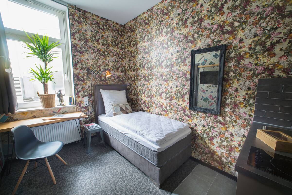 Un dormitorio con cama, escritorio, silla, radiador, ventana y planta en maceta.