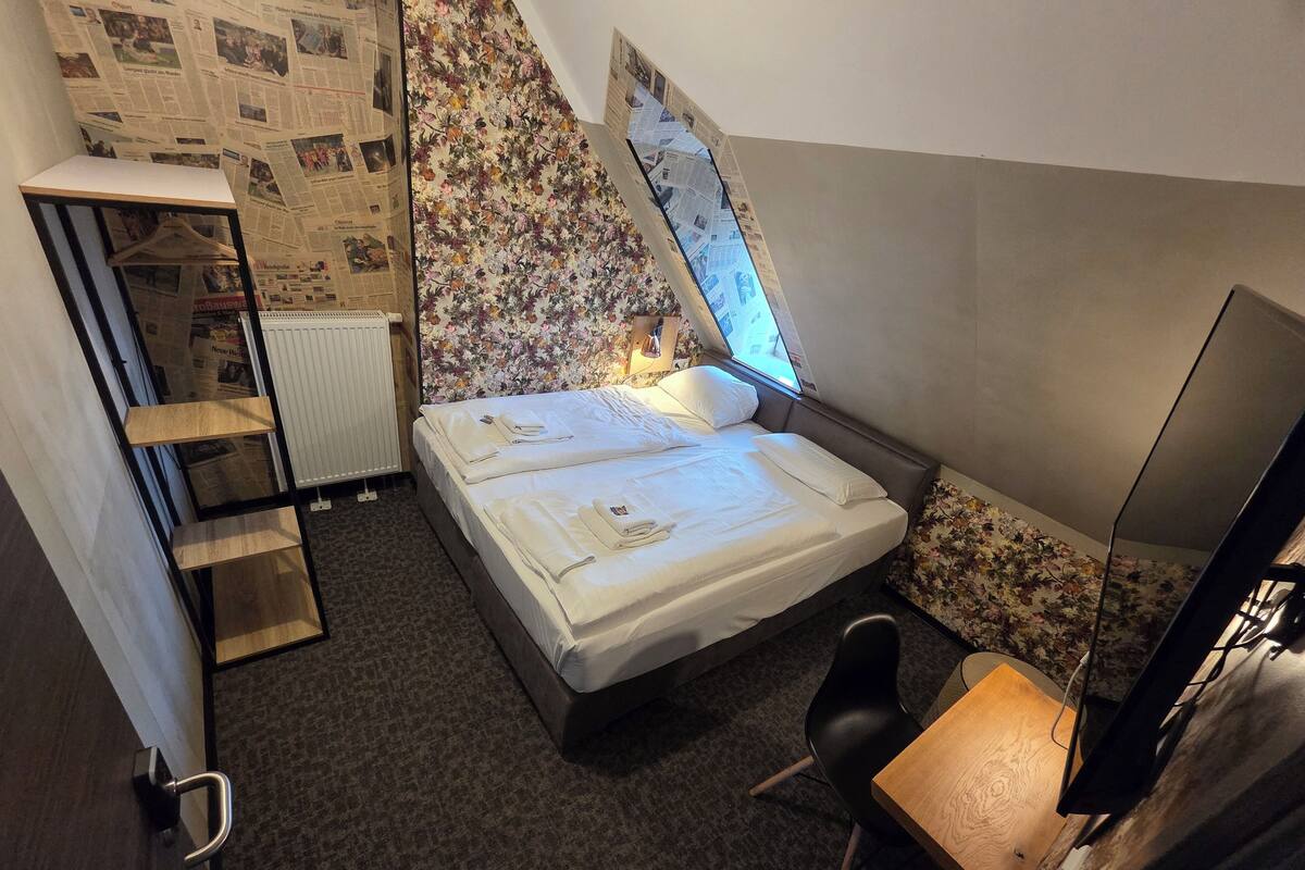 Habitación pequeña y funcional con cama, escritorio y silla. Hay un radiador y una ventana con vistas al exterior.