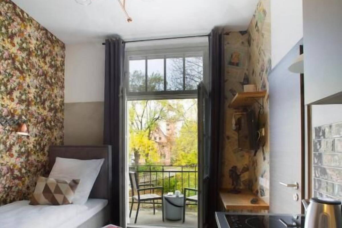 Un apartamento moderno con cama, un taburete pequeño, una cocina con lavabo y un balcón con vistas a árboles y edificios.