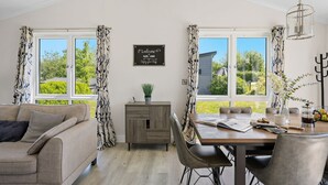 A cozy room with a sofa, a wooden dining table, and two windows with curtains. A sign on the wall welcomes guests to the lodge. - Hazel Lodge, 27 Roadford Lake Lodges (Lifton)